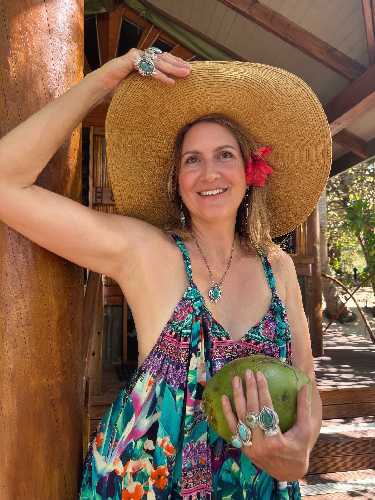 Woman wearing a wide-brimmed hat and turquoise jewelry, holding a green coconut, standing in a tropical setting.