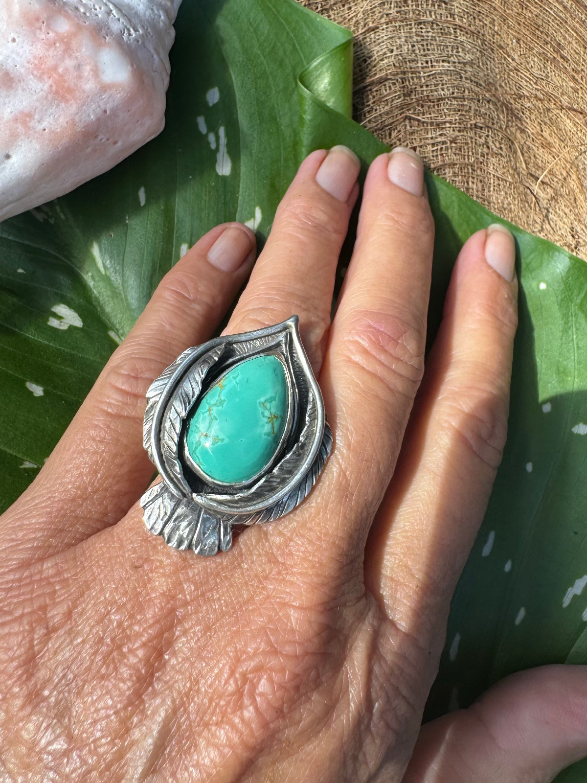Hand wearing a silver stylized bird ring with a turquoise stone on a green leaf background