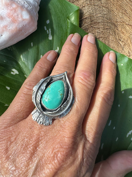 Hand wearing a silver stylized bird ring with a turquoise stone on a green leaf background