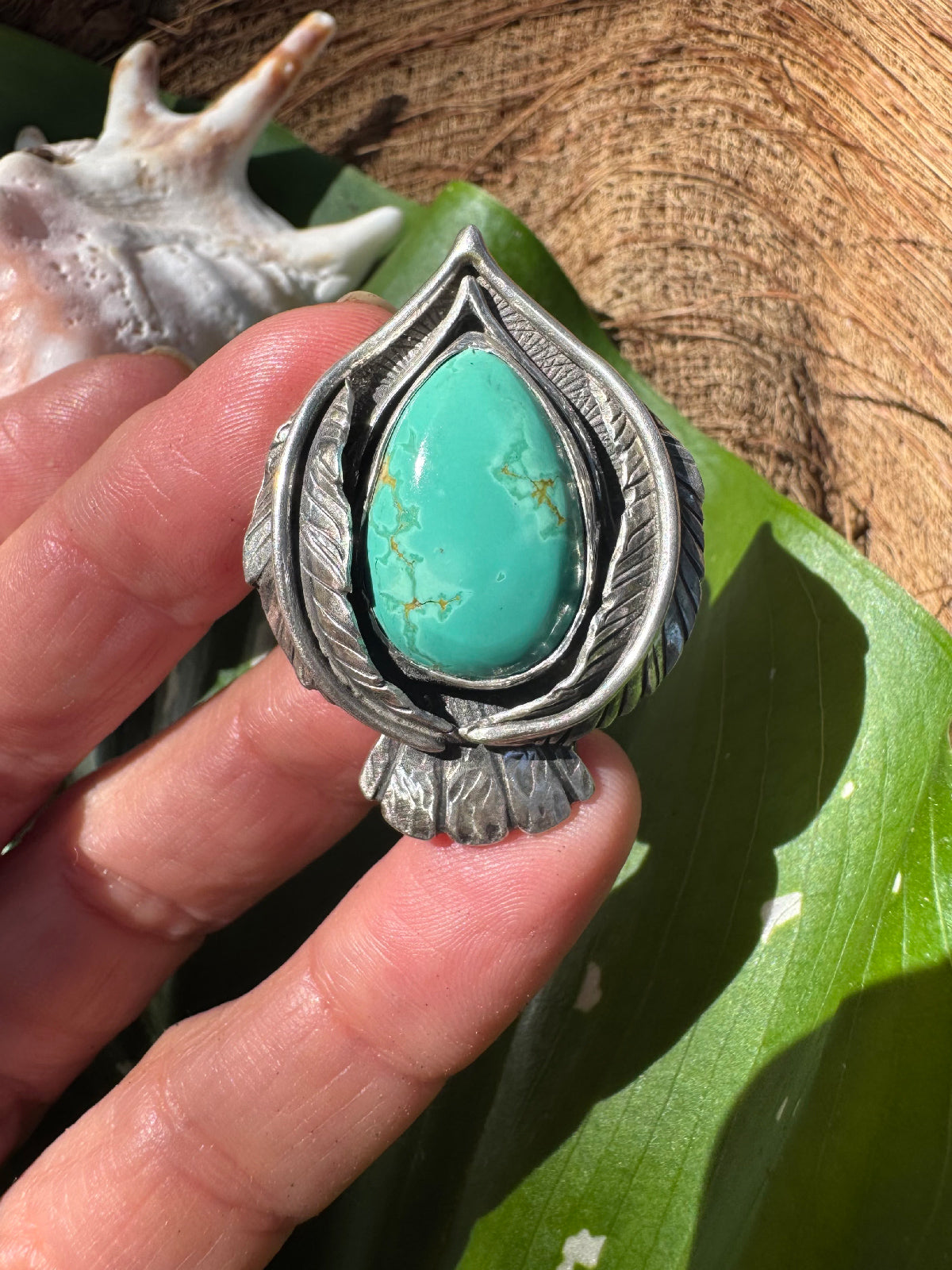 Turquoise stone in a silver stylized bird setting held by a hand on a leafy background