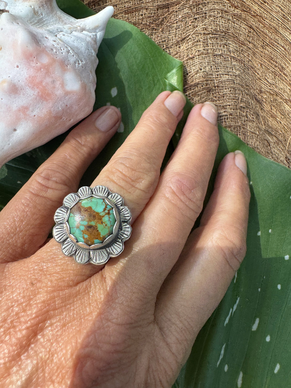 Hand wearing a silver flower ring with a turquoise stone on a leafy background