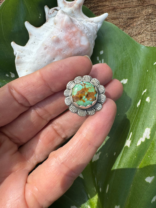 Silver flower ring with a turquoise stone held in a hand, with a shell and leaf in the background.
