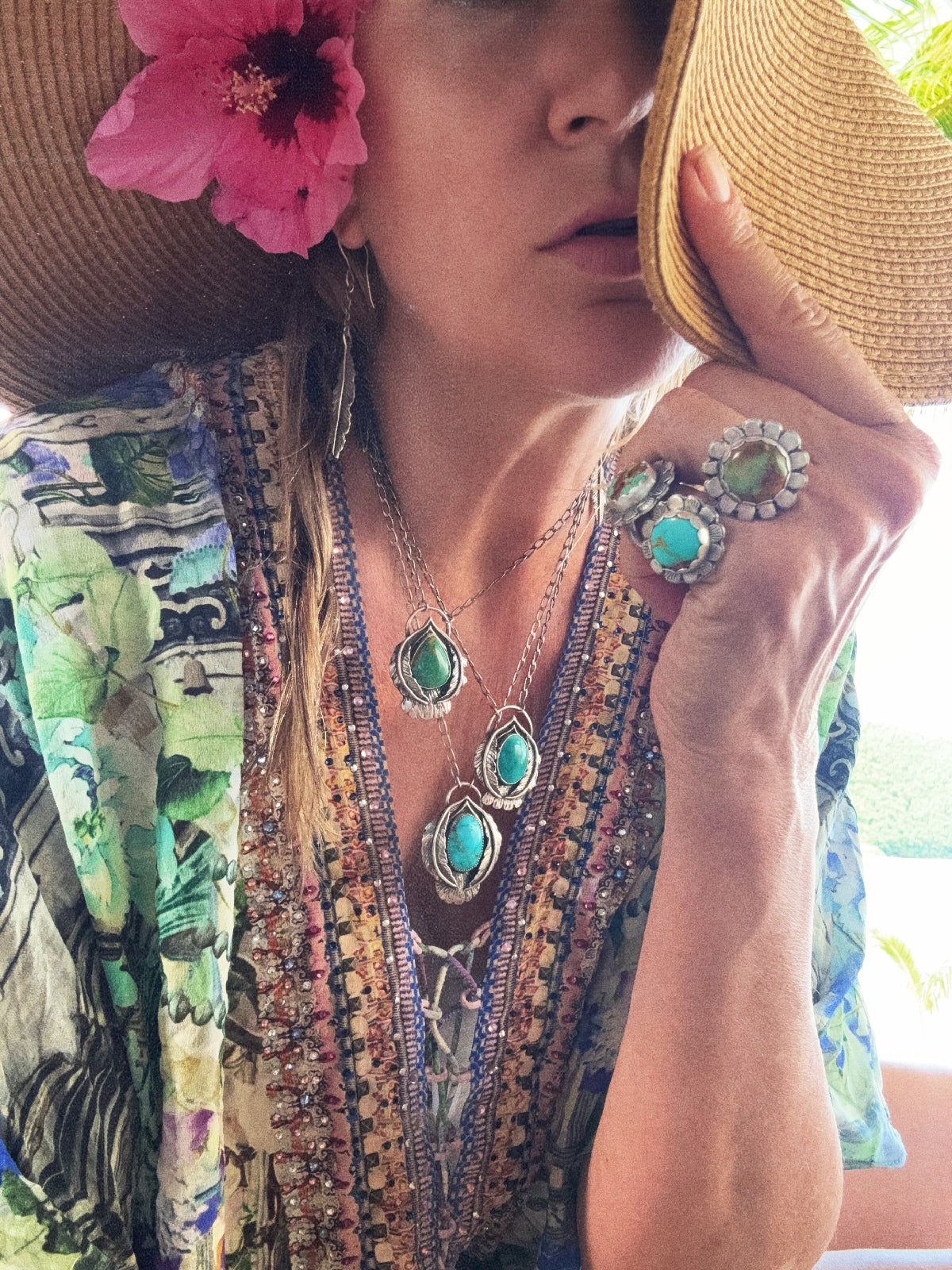 Woman wearing a colorful patterned top, straw hat, and turquoise jewelry outdoors.