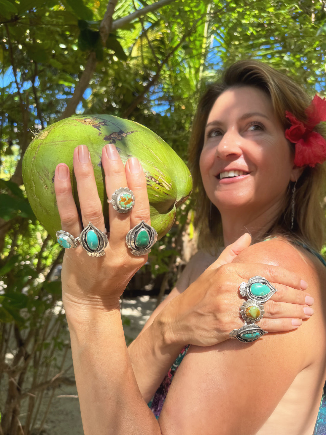 Woman holding a green coconut with turquoise rings on her fingers, surrounded by foliage.