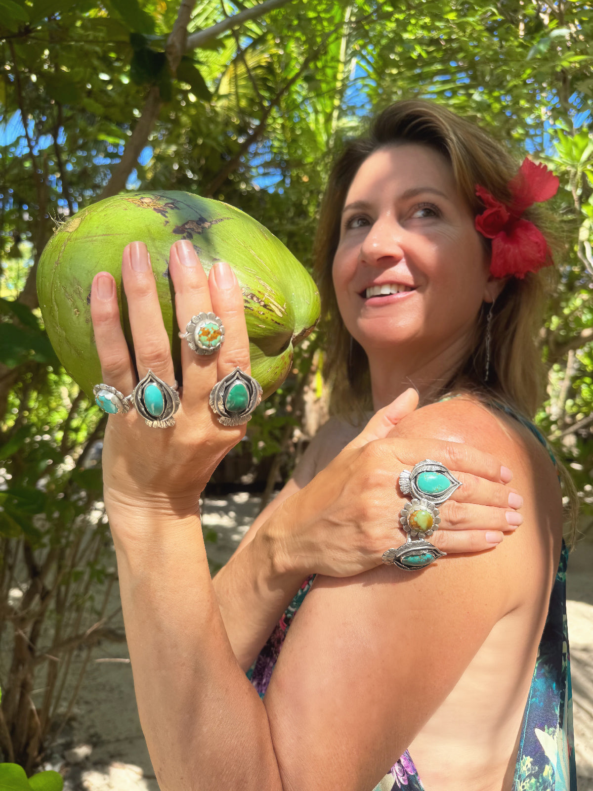 Woman holding a green coconut with turquoise rings on her fingers, surrounded by foliage.