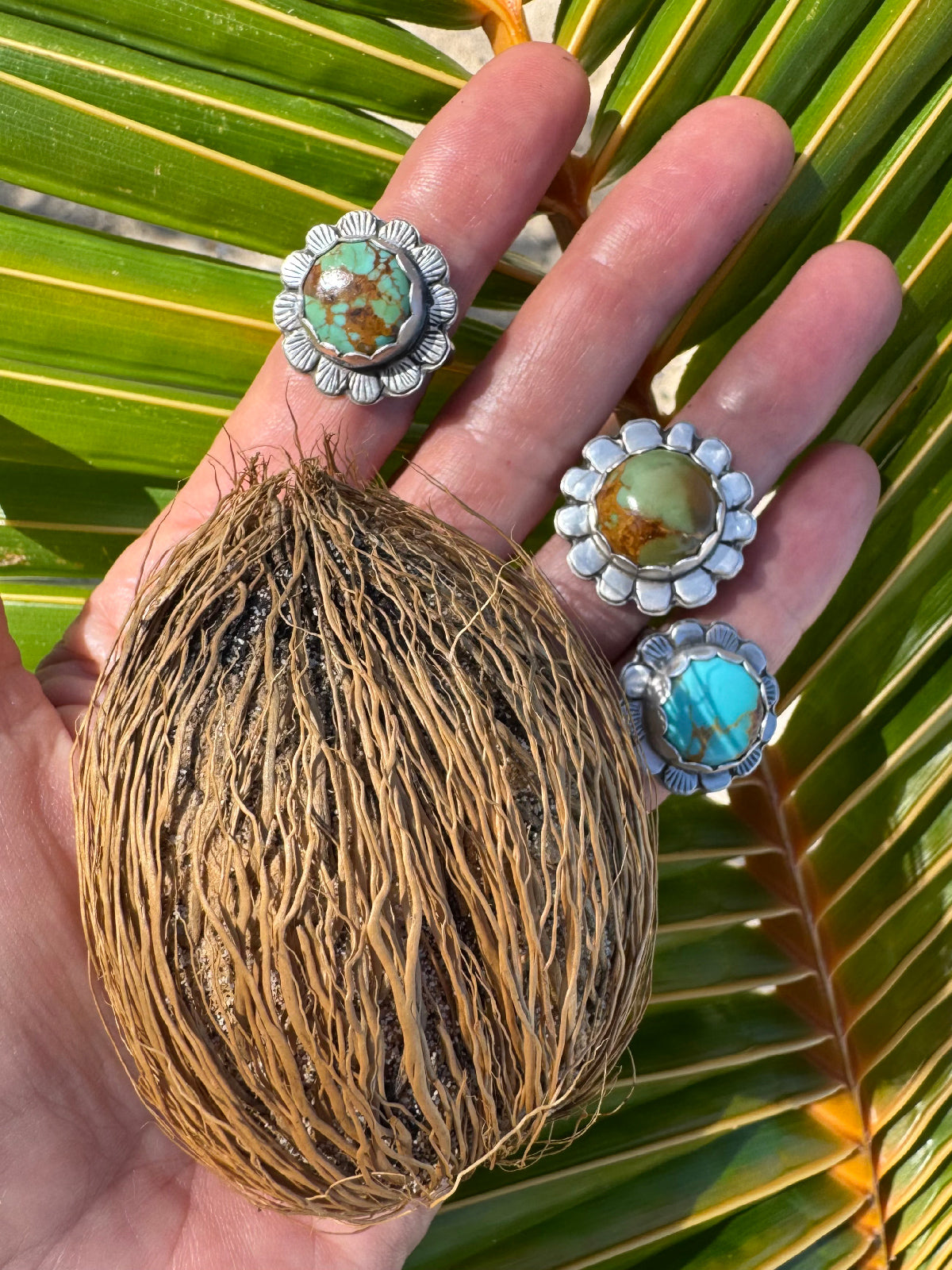 Hand holding a coconut with turquoise flower rings against a palm leaf background